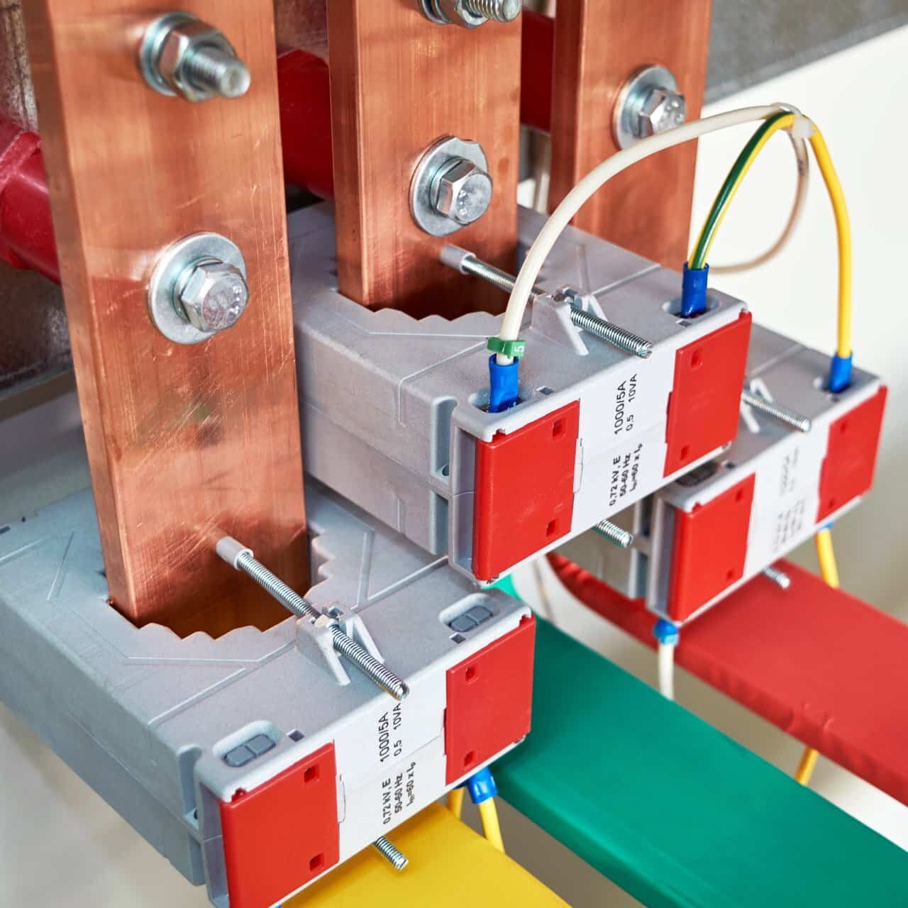 Electrical current transformers fitted on vertical copper busbars. Electrical wires are connected to the transformers. Copper tires are color-coded. The equipment is fixed in an electric Cabinet.